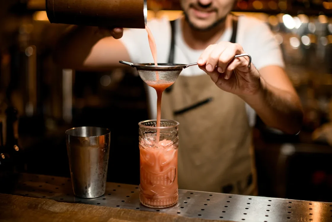 Bartender expertly strains vibrant cocktail into elegant glass at a cozy bar.