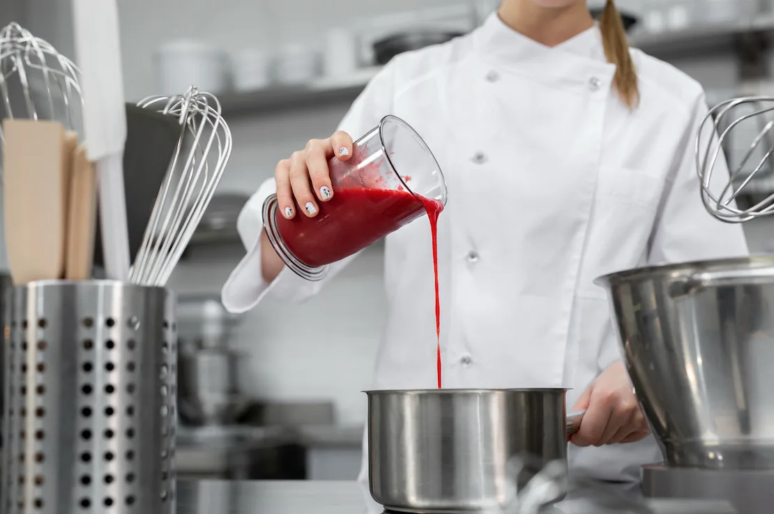 Chef pouring vibrant red sauce into a stainless steel pot in a busy kitchen.
