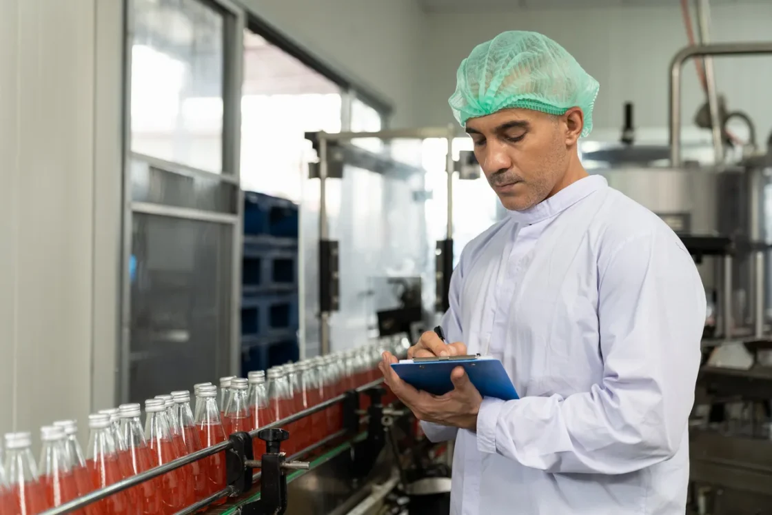 Male worker in hairnet inspecting basil seed beverage on conveyor belt in factory.